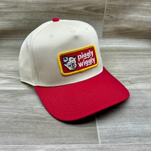 以下が含まれることがあります： 「Piggly Wiggly」と書いてある黄色と赤のパッチが付いた、ベージュと赤のベースボールキャップ。パッチには、漫画の豚が描かれている。