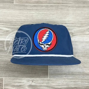 Könnte beinhalten: Marineblaue Kappe mit weißem Seildetail und flachem Schirm. Die Kappe hat einen runden Aufnäher mit einem Totenkopf- und Blitzmotiv in Rot, Blau und Weiß. Die Kappe ist im Retro-Stil.