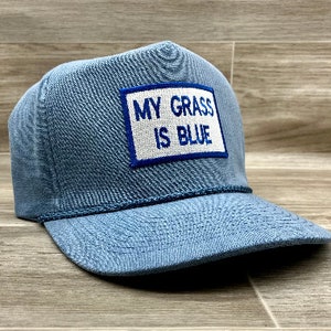 以下が含まれることがあります： 青いデニムのトラッカーハットに、白いパッチが縫い付けられており、青い文字で「MY GRASS IS BLUE」と書かれています。