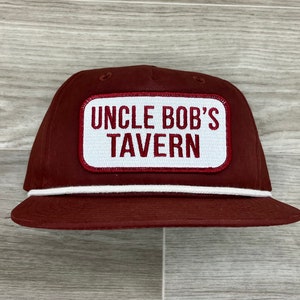May include: A maroon trucker hat with a white rope trim and a white patch that says "Uncle Bob's Tavern" in red letters.
