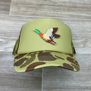May include: A green camouflage trucker hat with a brown, green, and blue embroidered duck flying on the front.