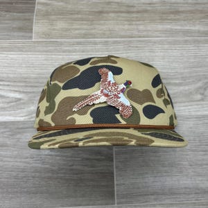Puede incluir: Una gorra de béisbol de camuflaje con una visera marrón y un faisán bordado en la parte delantera.