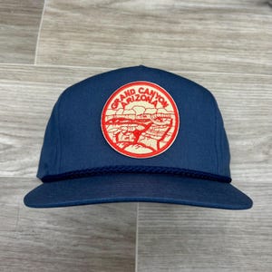 Puede incluir: Gorra de béisbol azul marino con un parche circular con una ilustración roja y crema del Gran Cañón. El parche está centrado en la parte delantera de la gorra, con las palabras "Grand Canyon Arizona" en un diseño circular.