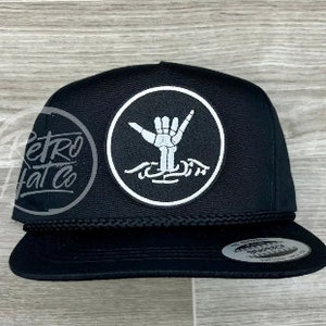 May include: Black flat-brimmed hat with a white circular patch featuring a skeleton hand in the shaka sign. The hat has a black rope detail and a curved brim. The text "Retro Hat Co" is on the side.