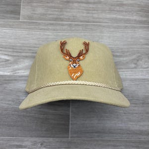 May include: Tan corduroy baseball cap with a detailed embroidered deer head patch. The deer has brown antlers and an orange and white face. A braided rope trim accents the brim.