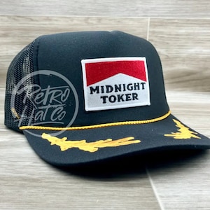May include: Black trucker hat with a yellow rope trim and a gold embroidered design on the brim. The hat features a white patch with red and white lettering that reads "MIDNIGHT TOKER".