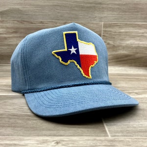May include: A blue cap with the Texas state flag embroidered on the front. The flag design features a white star on a blue background, a white and red stripe, and a yellow border. The cap has a rope detail and a curved peak.