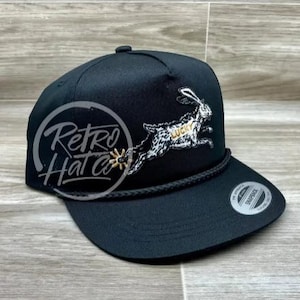Peut inclure: Casquette de baseball noire avec une visière plate et un détail de corde. La casquette présente un motif brodé d'un lapin courant avec le mot "LUCKY" et un éperon. Le texte "Retro Hat Co" est également visible.