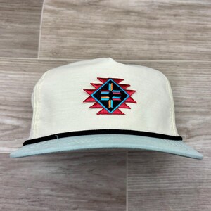 May include: A cream-colored baseball cap featuring a central embroidered geometric design in red, black, and light blue. The cap includes a thin black cord trim separating the crown from the light blue brim, creating a distinct contrast.
