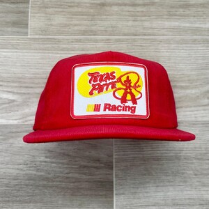 Puede incluir: Gorra roja de pana con visera plana. El parche rectangular muestra "Texas Pete Racing" en amarillo, rojo y blanco. El diseño incluye un vaquero y una cuerda.