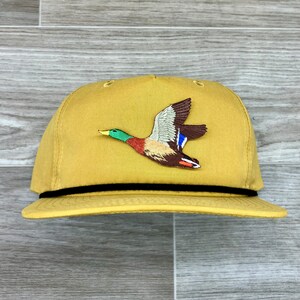 Puede incluir: Una gorra de béisbol amarilla con una banda negra y un pato bordado marrón y verde volando en la parte delantera.