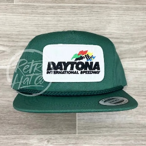 May include: A green baseball cap with a white patch featuring the Daytona International Speedway logo. The logo is a colorful checkered flag with the words "DAYTONA INTERNATIONAL SPEEDWAY" underneath.