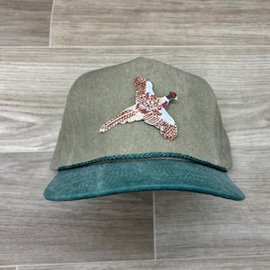 Może przedstawiać: Zielono-brązowa czapka z daszkiem z haftowanym wzorem bażanta. Czapka ma zielony daszek i brązową koronę.