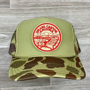 May include: A green and brown camouflage trucker hat with a red and white embroidered patch that says "Grand Canyon Arizona".