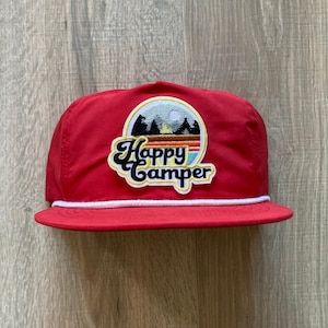 May include: Red baseball cap with a white rope trim and a white patch featuring a retro-style graphic that says "Happy Camper" and depicts a mountain scene with a campfire and a full moon.
