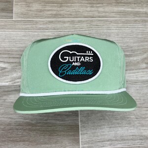 May include: A light green baseball cap with a white brim and a black and white patch that says "Guitars and Cadillacs" in a cursive font.