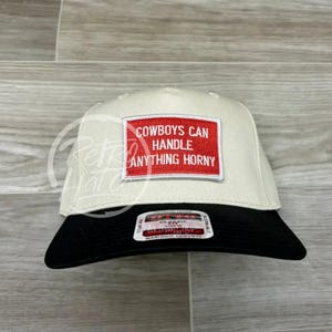 Peut inclure: Une casquette de baseball blanche avec une visière noire. Le devant de la casquette a un patch rouge avec le texte "COWBOYS CAN HANDLE ANYTHING HORNY".