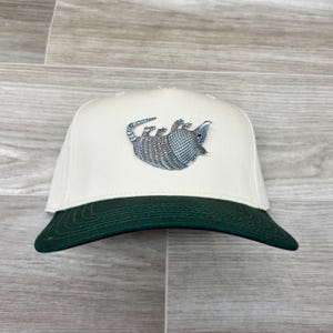 May include: A white and green baseball cap with an embroidered armadillo design. The armadillo is facing the left side of the cap.