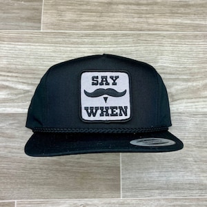 Puede incluir: Gorra de camionero negra con un parche cuadrado blanco que dice "SAY WHEN" con un bigote negro sobre el texto.
