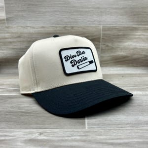 May include: A beige and black trucker hat with a white patch that says "Dive Bar Darlin" with a cigarette graphic.