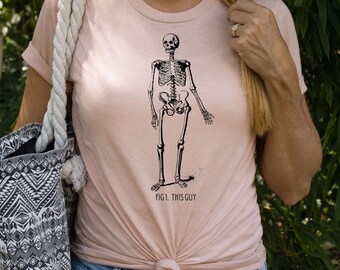 Può includere: T-shirt rosa chiaro con un'illustrazione in bianco e nero di uno scheletro umano. Il testo "FIG. 1. THIS GUY" è stampato sotto. La maglietta è annodata in vita. È visibile una borsa tote con un motivo in bianco e nero.