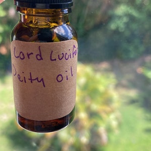 May include: A small brown glass bottle with a black dropper top. The bottle has a brown paper label with the text "Lord Lucifer Deity Oil" written in purple ink.