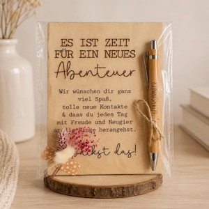 Könnte beinhalten: Eine rechteckige Holztafel in transparenter Folie mit dem deutschen Text ES IST ZEIT FÜR EIN NEUES Abenteuer, einem weiteren Text und dem Satz Du packst das! Ein Bambuskugelschreiber ist mit einer Kordel befestigt, und unten links befindet sich ein kleines Trockenblumenarrangement.