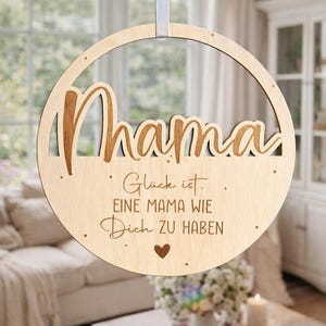 Könnte beinhalten: Runder Holzschild mit dem Wort "Mama" in kursiver Schrift ausgeschnitten. Darunter befindet sich gravierter deutscher Text und ein kleines Herz. Ein graues Band ist zum Aufhängen befestigt.