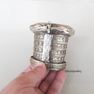 Può includere: Un bracciale a polsino argentato con intricati motivi geometrici e una grande pietra ovale marrone-rossastra. Il bracciale ha un design a cerniera ed è tenuto aperto. Gioielli vintage o antichi.