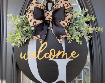Op de afbeelding: Een rond zwart welkom bord met een witte letter G en gouden letters die "welcome" zeggen. Het bord is versierd met een luipaardprint strik en groen.