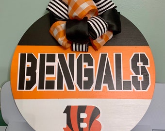 May include: A round wooden sign with the word "BENGALS" in black letters on an orange background. The sign also features a black and orange striped bow and a black and orange "1B" logo.