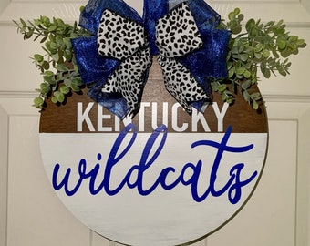 May include: A round wooden door hanger with a white and brown background. The words "Kentucky Wildcats" are written in blue cursive lettering. A blue and white leopard print bow is attached to the top of the hanger.