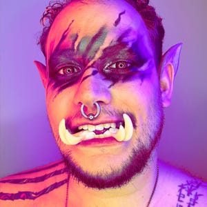 May include: A person with a shaved head and a beard is wearing a costume with a large, white, fake teeth, a nose ring, and black and green face paint. The person is wearing a silver chain necklace and has a tattoo on their chest.