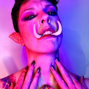 May include: A person with short dark hair and bright pink and blue makeup, wearing white tusks and dark nail polish. The person is looking at the camera with a serious expression.