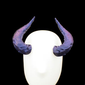 Puede incluir: Un par de cuernos curvos y texturizados en tonos morados y azules, montados sobre una forma de cabeza blanca de forma ovalada. Los cuernos tienen una superficie detallada y estriada, sobre un fondo negro.