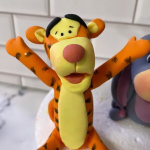 May include: Orange and yellow fondant figurine of Tigger from Winnie the Pooh, with black stripes and a big smile. The figurine is sitting on a white cake.