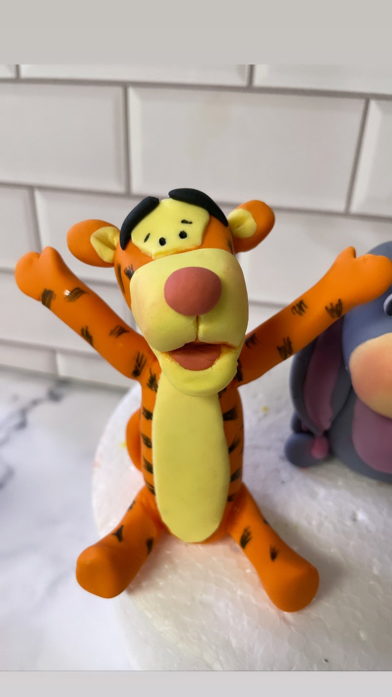 May include: Orange and yellow fondant figurine of Tigger from Winnie the Pooh, with black stripes and a big smile. The figurine is sitting on a white cake.