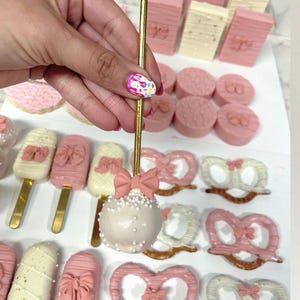 May include: A collection of pink and white treats, including cake pops, cookies, and pretzels. The cake pops are decorated with white sprinkles and a pink bow. Other treats include pink and white chocolate-covered pretzels and cookies with bow designs.
