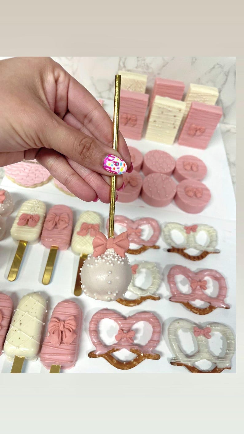 May include: A collection of pink and white treats, including cake pops, cookies, and pretzels. The cake pops are decorated with white sprinkles and a pink bow. Other treats include pink and white chocolate-covered pretzels and cookies with bow designs.