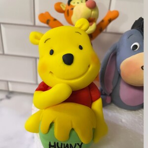 May include: A yellow Winnie the Pooh figurine with a red shirt, holding a green pot with the word "Hunny" written on it. The figurine is part of a set of three, including Tigger and Eeyore.