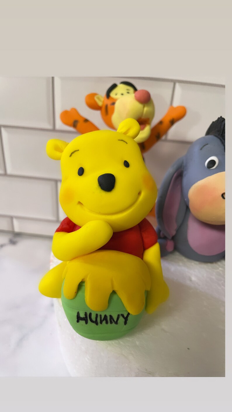 May include: A yellow Winnie the Pooh figurine with a red shirt, holding a green pot with the word "Hunny" written on it. The figurine is part of a set of three, including Tigger and Eeyore.