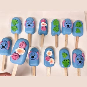 May include: A collection of blue, popsicle-shaped treats on wooden sticks. Some are decorated with a cartoon character face, others with flowers or green leaves. The treats are arranged in rows, offering a colorful and playful aesthetic.