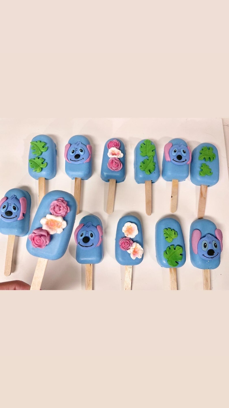 May include: A collection of blue, popsicle-shaped treats on wooden sticks. Some are decorated with a cartoon character face, others with flowers or green leaves. The treats are arranged in rows, offering a colorful and playful aesthetic.