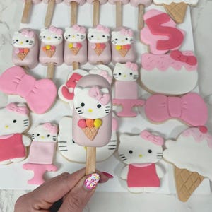 May include: A collection of pink and white Hello Kitty-themed treats, including cake pops, cookies, and ice cream cone-shaped desserts. The cake pops feature Hello Kitty faces with ice cream cone details. The cookies are decorated with bows and the number 5.