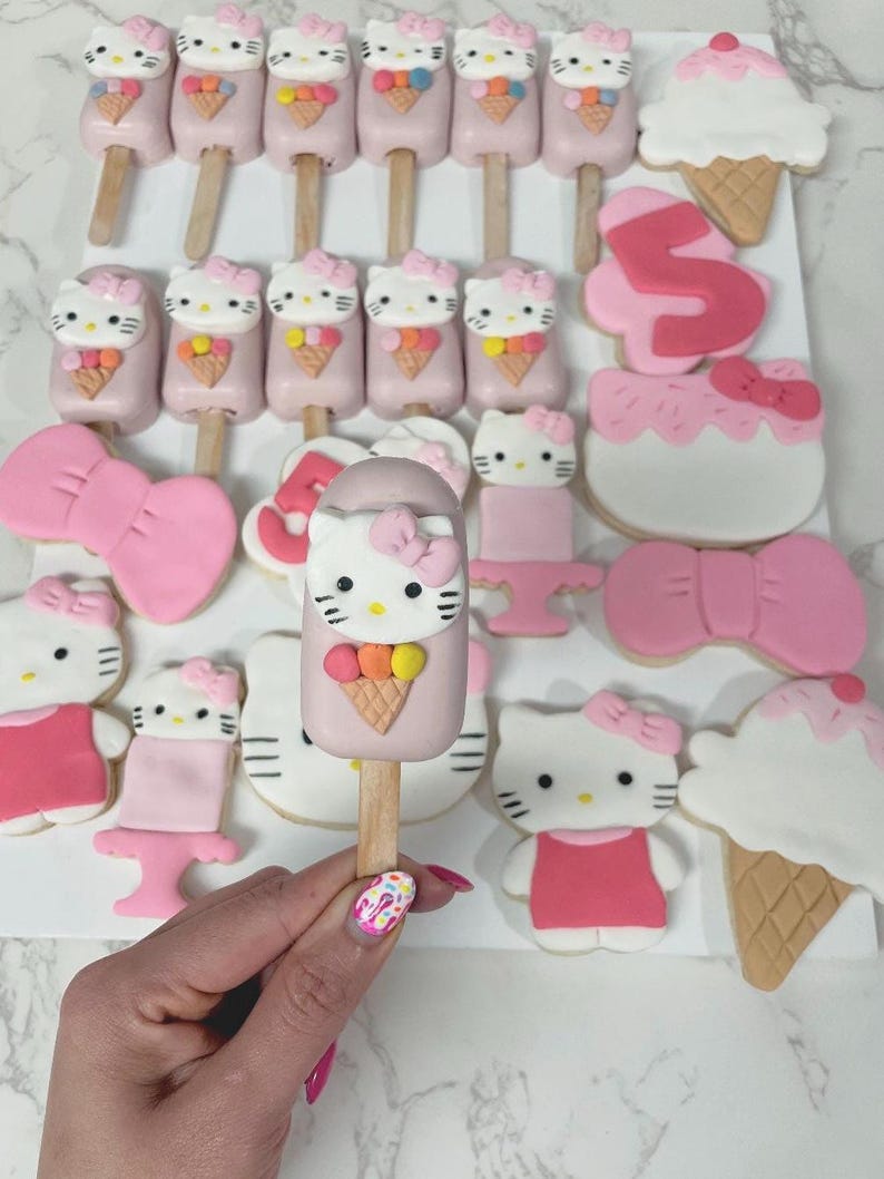 May include: A collection of pink and white Hello Kitty-themed treats, including cake pops, cookies, and ice cream cone-shaped desserts. The cake pops feature Hello Kitty faces with ice cream cone details. The cookies are decorated with bows and the number 5.