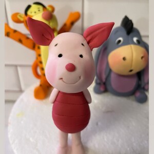 May include: A set of three Winnie the Pooh character cake toppers. The toppers are made of fondant and feature Piglet, Tigger, and Eeyore. Piglet is pink with a red shirt, Tigger is orange and black with yellow stripes, and Eeyore is gray with a purple belly.