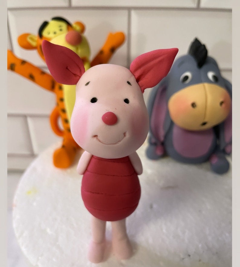 May include: A set of three Winnie the Pooh character cake toppers. The toppers are made of fondant and feature Piglet, Tigger, and Eeyore. Piglet is pink with a red shirt, Tigger is orange and black with yellow stripes, and Eeyore is gray with a purple belly.