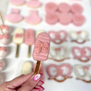 May include: Pink-colored treats, including a popsicle-shaped cake pop with a bow design, cookies, and pretzels. The cake pop is held by a person with a gold stick. Other treats include baby bootie cookies and heart-shaped pretzels.