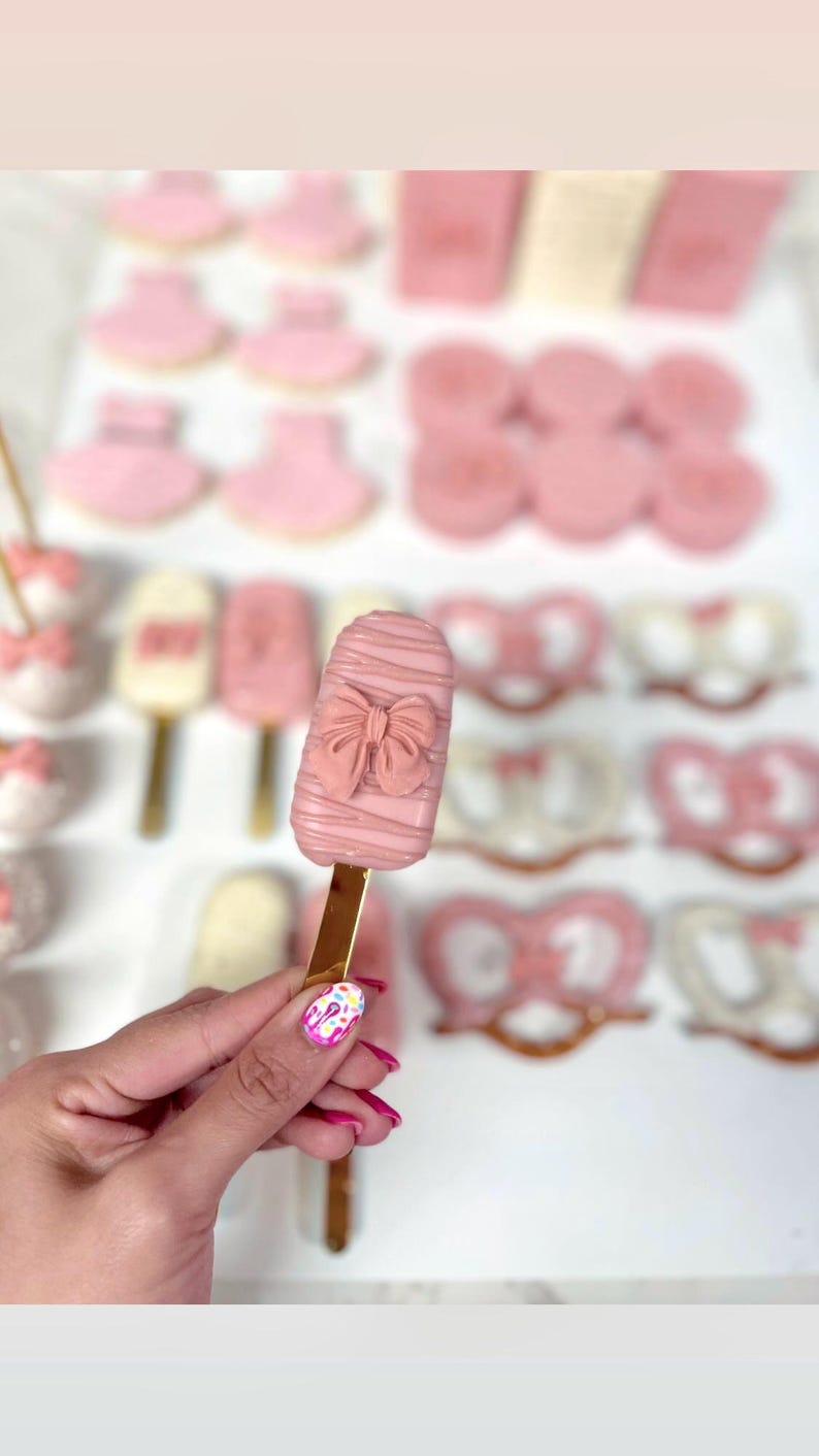 May include: Pink-colored treats, including a popsicle-shaped cake pop with a bow design, cookies, and pretzels. The cake pop is held by a person with a gold stick. Other treats include baby bootie cookies and heart-shaped pretzels.