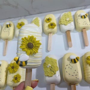 May include: A collection of decorative, bee-themed treats on wooden sticks. The treats are yellow and white, decorated with yellow flowers, honeycomb patterns, and small bee figures. The treats are arranged in rows.